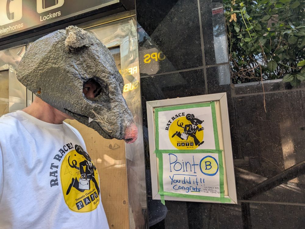 A racer in a rat mask beside a hand-lettered 'Point B, you did it' sign
