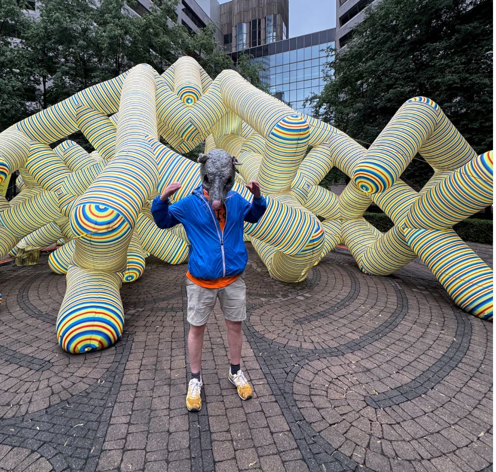 A racer in a rat mask in front of a giant striped public sculpture